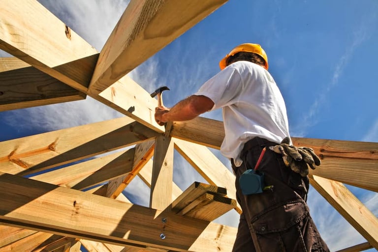 Construction worker in safety gear installing wooden roof framing on a building site under blue sky